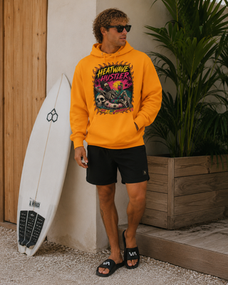 A man wearing a bright orange graphic hoodie with 'Heatwave Mystery' printed on the front, black shorts, and black slides, standing next to a white surfboard in a beach-themed setting.